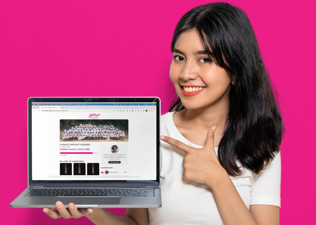 A young woman holding a computer in one hand and pointing to the screen on the other. On the screen is LipRevolt's fundraising page.
