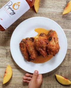 Chicken wings on a white plate sitting on a wood table surrounded by peach slices and a bottle of Wing Suite Piping Peach hot sauce