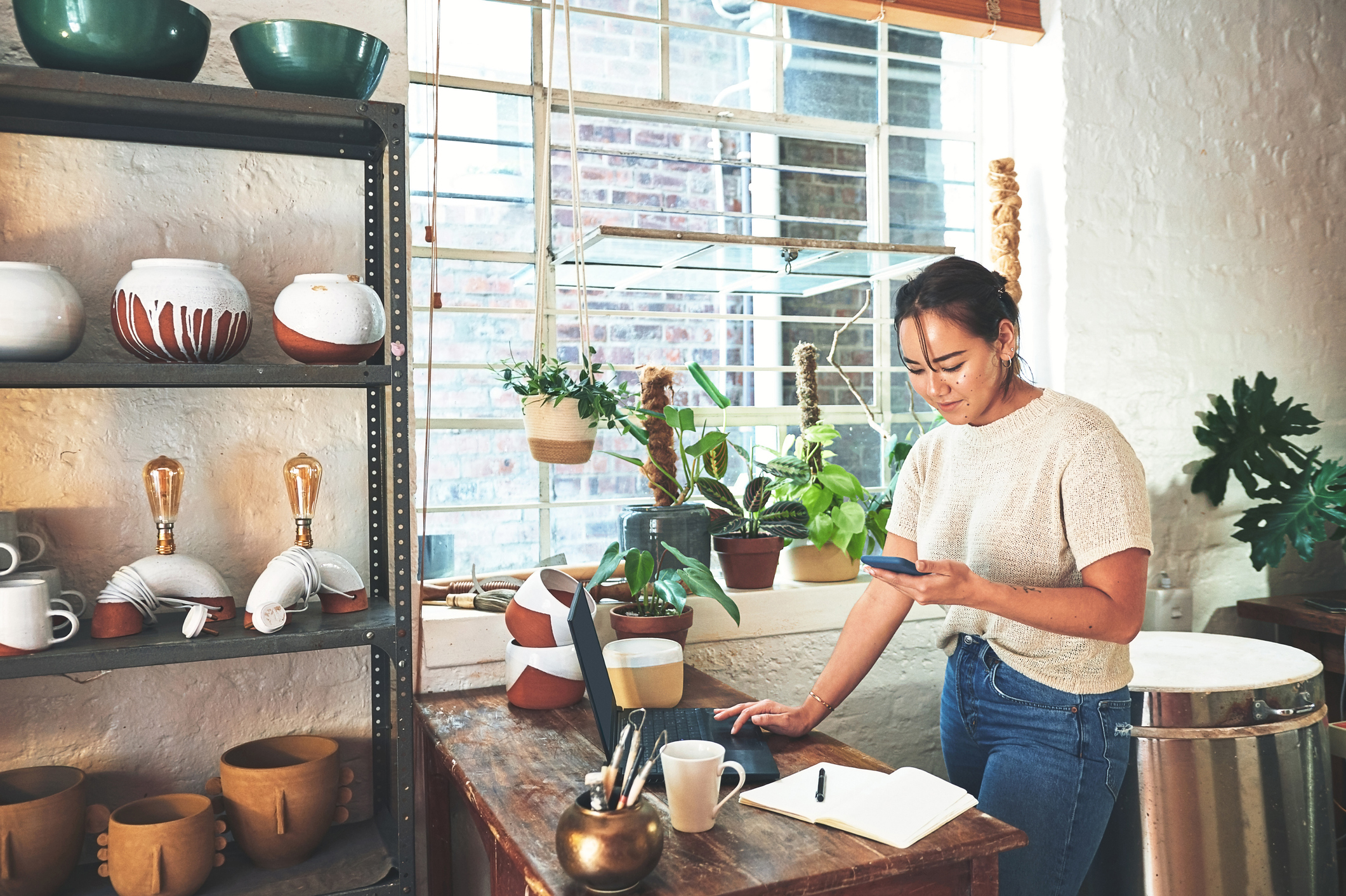 Cropped shot of an attractive young business owner standing and using her laptop and cellphone in her potter