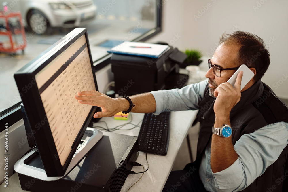 A man multitasks at his desk, pointing at data on a computer screen while speaking on his phone.