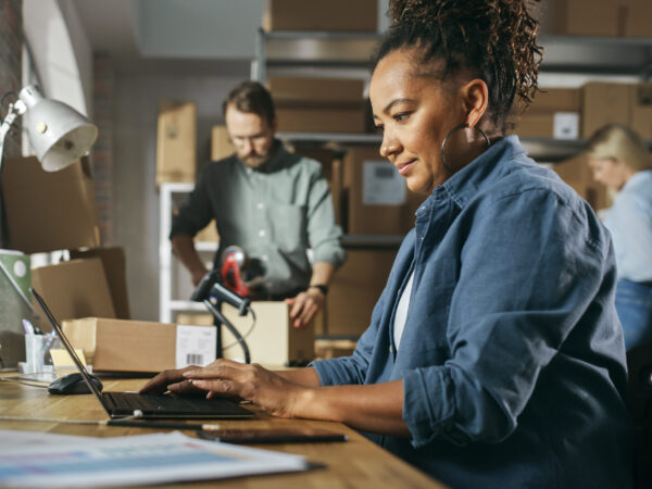 Inventory Manager Using Laptop Computer to Check Order Number on a Parcel, Preparing a Small Cardboard Box for Postage.